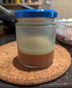 A small glass jar with reserved chicken drippings. It shows a layer of solid fat on top of dark golden jelled chicken juices.