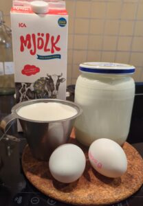 A stainless measuring cup of buttermilk sits next to the glass jar it came from. Two eggs sit in front, and a carton of whole milk behind them.