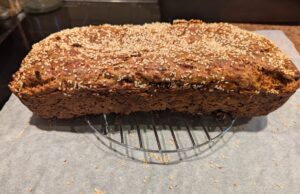 Bare loaf of bread sitting on a raised round wire rack, above a sheet of white parchment paper scattered with sesame seeds and crumbs