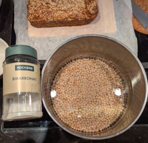 Dry peas soaking in a pot of water, with a jar of sodium bicarbonate laid on one of the handles.