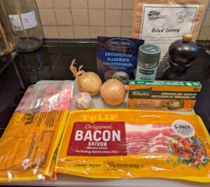A collection of ingredients, including packaged bacon, a box of vegetable bouillon cubes, two onions, a small head of garlic, and containers of dried summer savory, marjoram, and whole allspice.