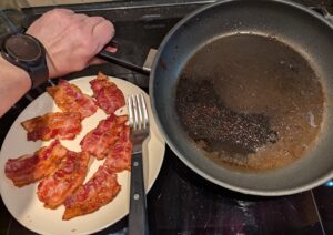 A small plate of freshly cooked crispy streaky bacon slices with the turning fork resting on the edge. To the right of this, I tilt the bacon cooking skillet to show the browned residue and grease left in there from it.
