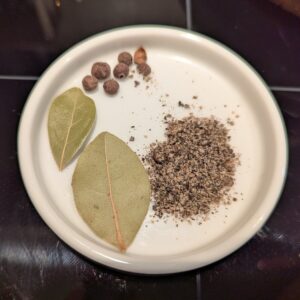 A small shallow ceramic dish holds bay leaves, whole allspice, and a pile of ground pepper