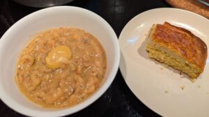 A bowl of yellow pea soup topped with a dollop of brown mustard in the center sits to the left of a side plate holding a piece of cornbread.