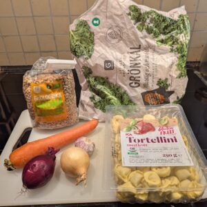 Major ingredients laid out on a white cutting board, including a bag of lentils, a carrot, two small onions, a head of garlic, a partial bag of frozen chopped kale, and a package of fresh tortellini