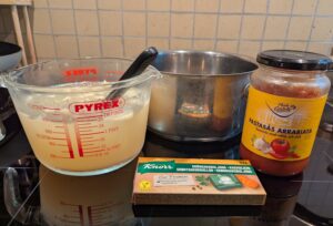 A large Pyrex measuring cup of homemade chicken broth sits next to a mostly full jar of Arrabiata pasta sauce and a box of Knorr vegetable bouillon cubes. Behind it all sits the intended soup pot.