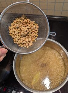 A metal mesh strainer of washed lentils poised to pour into a pot of broth with the initial seasonings floating on the surface.