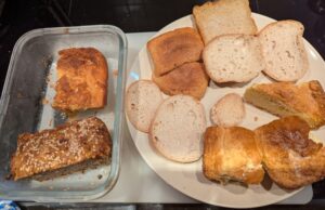 A collection of sliced bread heels and pieces from previous batches of homemade cornbread, with the frozen pieces laid out on a dinner plate to thaw. To the side sits a glass container with another piece of cornbread and the end chunk from a recent loaf of quickbread posted on this blog, not frozen.