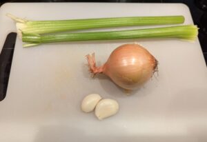 Two stalks of celery lie on a cutting board, above an onion and two peeled cloves of garlic.