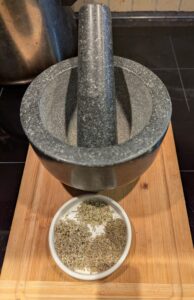 Dried herbs sit on a shallow ceramic dish, in front of a granite mortar and pestle.