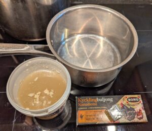 An open plastic container of chicken broth sits next to a box of chicken bouiilon cubes. A medium-sized saucepan sits behind them.