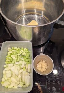 A plastic container of chopped onion and celery sits next to a small stainless measuring cup holding crushed garlic. Behind them sits a pot with butter visible inside.