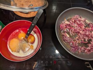 A bowl with eggs waiting to be scrambled, on a stovetop next to a pan of sautéing red onions
