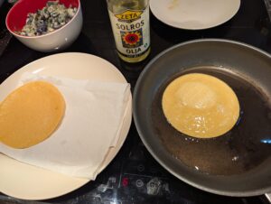 Tortilla frying in a pan, next to a paper towel lined plate with the previous cooked tortilla on it.