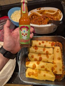 Rolled enchiladas lined up in the baking dish, with a smattering of hot sauce on top. A hand is also in the frame, holding a bottle to show the "Skånsk Chili Original Habanero Hot Sauce" bottle, with text surrounding a stylized sugar skull.
