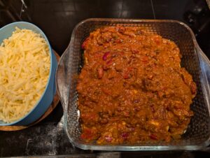 The same baking dish with a thick layer of chili spread over top of the rolled enchiladas, and a container of grated cheese ready to its left.