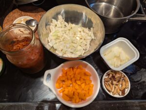 Clockwise from top left: A jar of shredded white cabbage kimchi, a bowl of fresh cabbage cut into small bite-sized pieces, a bowl of halved and sliced onion, a bowl of quartered and sliced carrots