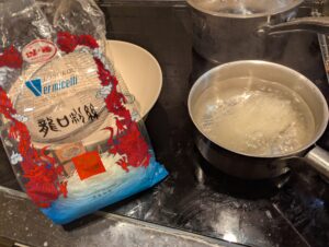 A small pan of boiling noodles, with the open package and a waiting soup bowl next to it