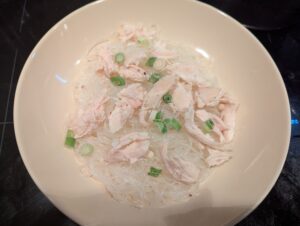 The soup bowl with hot noodles, topped with shreds of cooked chicken breast and a handful of frozen sliced green onion