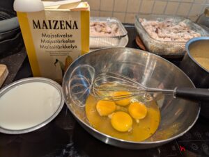 The same stainless mixing bowl that held the bread cubes earlier, with a wire whisk resting in it.. Now it contains four raw eggs removed from the shells. next to it sit a box of cornstarch and a measuring cup with milk.