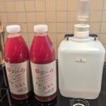 Two liter plastic bottles of a cloudy pinkish red mixed fruit juice sit next to a plastic water jug with an airlock stopper on top