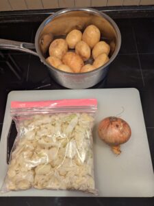 A zip bag of already cut cabbage sits with an onion and a pan with raw potatoes in it