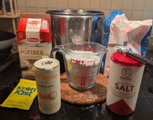 A measuring cup of flour, surrounded by packages of potato fiber, sugar, salt, xanthan gum, and dry yeast