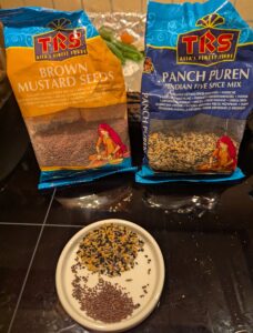 Bags of panch phoron seed mixture and dark mustard seeds, sitting in front of a small ceramic dish with seeds measured out.