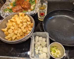The chopped potatoes, onion, garlic, and green chili sit in containers next to a nonstick frying pan and a bottle of sunflower oil