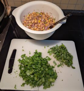 Chopped herbs on a cutting board, in front of the mixing bowl of salad in progress.