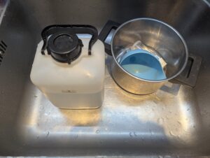 The fermentation jug sitting in a kitchen sink with a pot holding a plastic funnel and the jug plug with airlock.