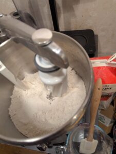 Dry ingredients in a bowl plaxed onto a stand mixer base with dough mixers visible. Beiside it sit a carton of extra flour, and a Pyrex measuring cup holding a rubber spatula ready..