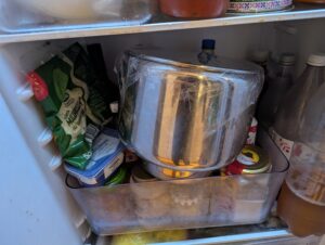 A large stainless steel mixer bowl sits in the bottom shelf of a rather crowded refrigerator.