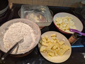 Mixed dough ingredients sit next to a plate of butter chunks and the sliced apples, with the baking dish in the background