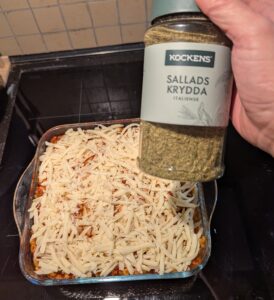 Assembled casserole with cheese on top, and a hand holding a bottle of Italian herb seasoning