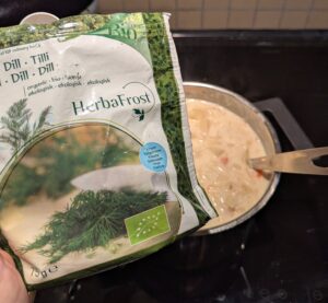 A bag of frozen chopped dill and measuring spoon held in front of the otherwise finished pot of soup