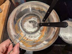 Lumpy powdered sugar being sifted into the icing bowl with a tea strainer and spoon