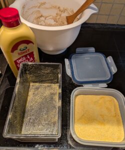 Greased bread pan lightly coated inside with cornmeal, next to the storage bowl of meal