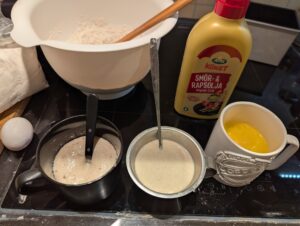 Wet ingredients in cups sit in front of the mixing bowl containing dry mixture and a big wooden spoon